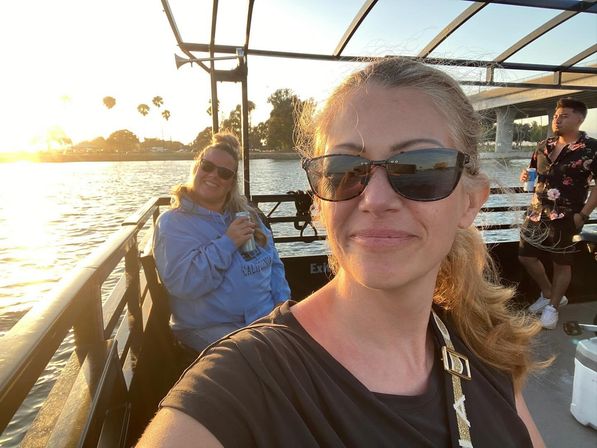 Friends on a sunset boat ride along a palm-lined waterfront, wearing sunglasses and holding drinks with a bridge in the background.