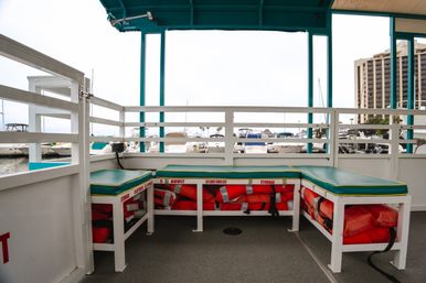 Covered boat deck with teal-cushioned benches and orange life jackets stowed under seats, marina with docked yachts visible beyond