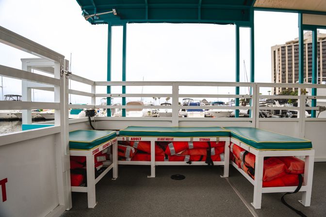 Covered boat deck with teal-cushioned benches and orange life jackets stowed under seats, marina with docked yachts visible beyond