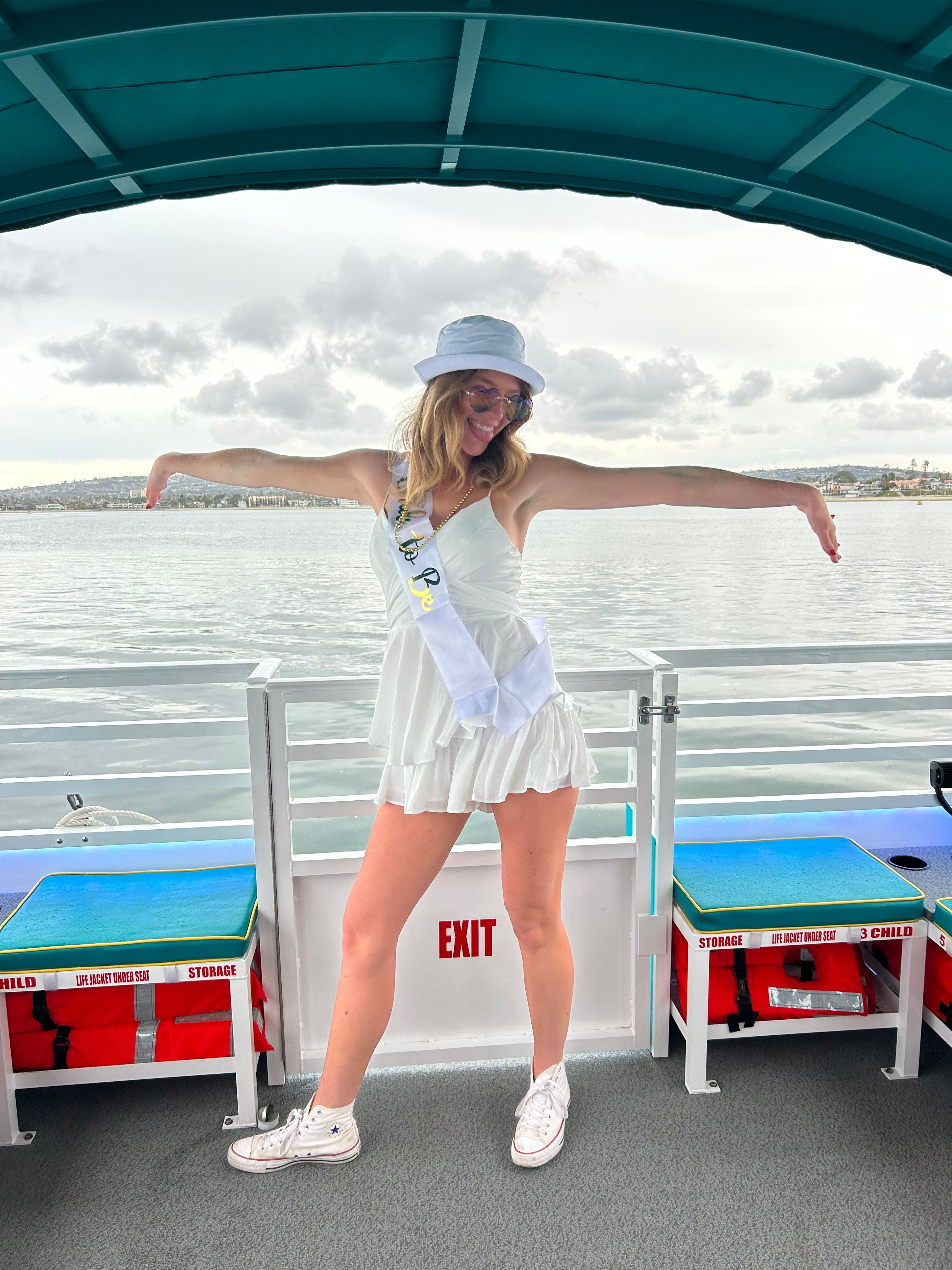 Woman in a white sundress, bucket hat and celebration sash striking a playful pose with arms outstretched on a covered party boat, calm bay water and cloudy coastal skyline in the background