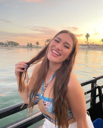 Smiling woman in a patterned bikini top and white shorts leaning on a boat railing, playing with her hair at a coastal harbor sunset with palm trees and calm water