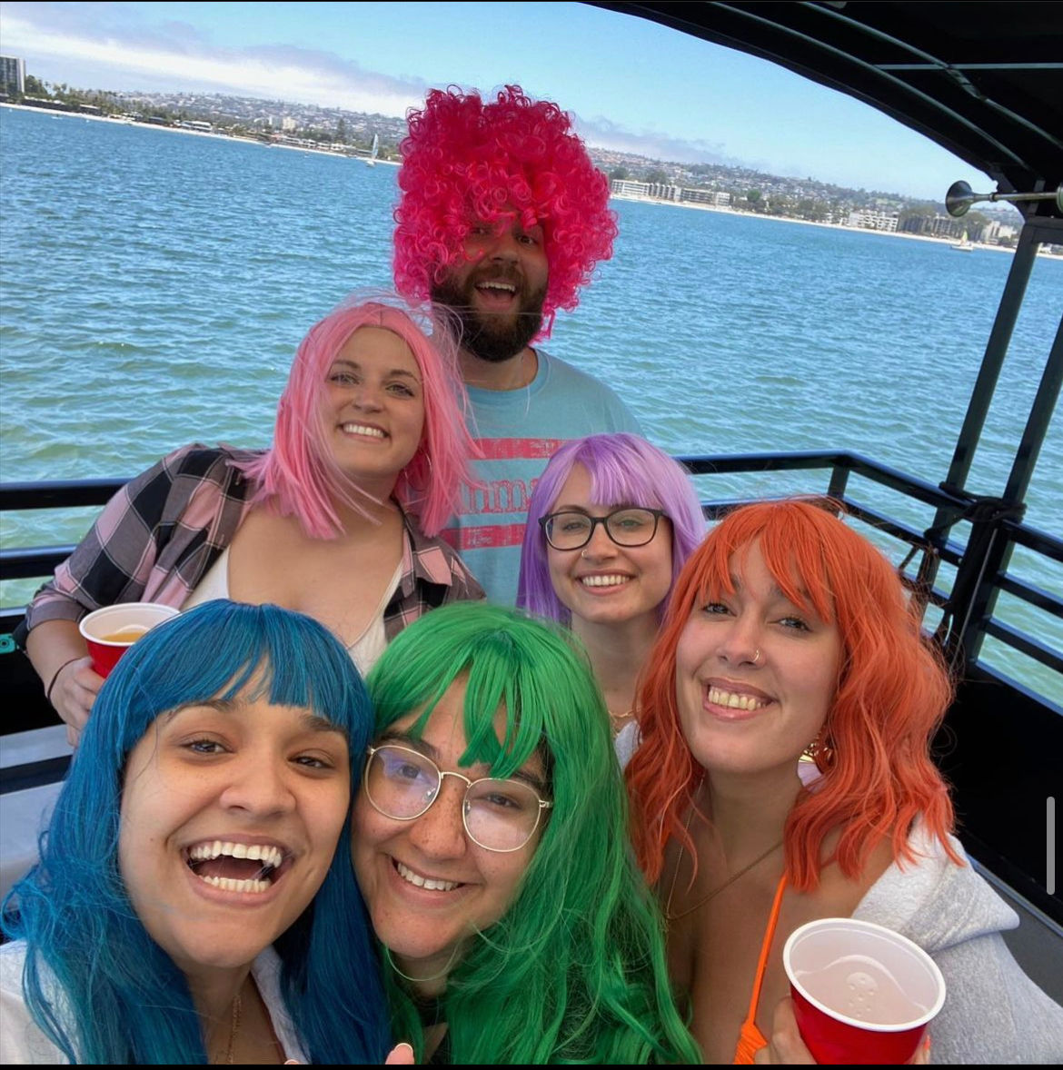 Smiling group of six friends on a boat party at a sunny bay, wearing colorful wigs and holding red cups.