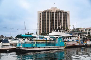 Turquoise pedal-powered party boat docked at a coastal marina with nearby yachts, a multi-story hotel tower in the background, string lights overhead and calm reflective water.