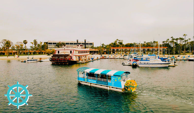Cheerful teal-and-white covered tour boat with a yellow paddlewheel cruising a palm-lined Southern California marina, with a riverboat-style paddlewheeler, docked sailboats and low-rise waterfront buildings in the background.