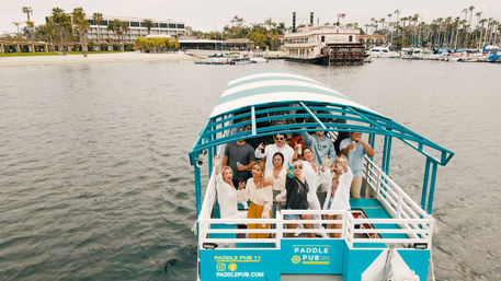 Group enjoying drinks on a turquoise covered paddle boat in a palm-lined coastal marina with hotels and docked yachts