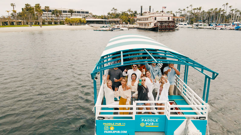 Group enjoying drinks on a turquoise covered paddle boat in a palm-lined coastal marina with hotels and docked yachts