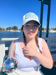 Smiling person with braided hair wearing a 'MARRIAGE MATERIAL' cap and white tank top, holding a mirrored disco-ball tumbler on a sunny boat at a marina with sailboats and blue sky