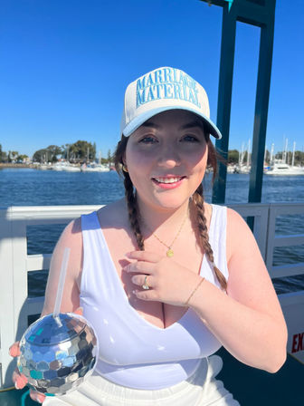 Smiling person with braided hair wearing a 'MARRIAGE MATERIAL' cap and white tank top, holding a mirrored disco-ball tumbler on a sunny boat at a marina with sailboats and blue sky