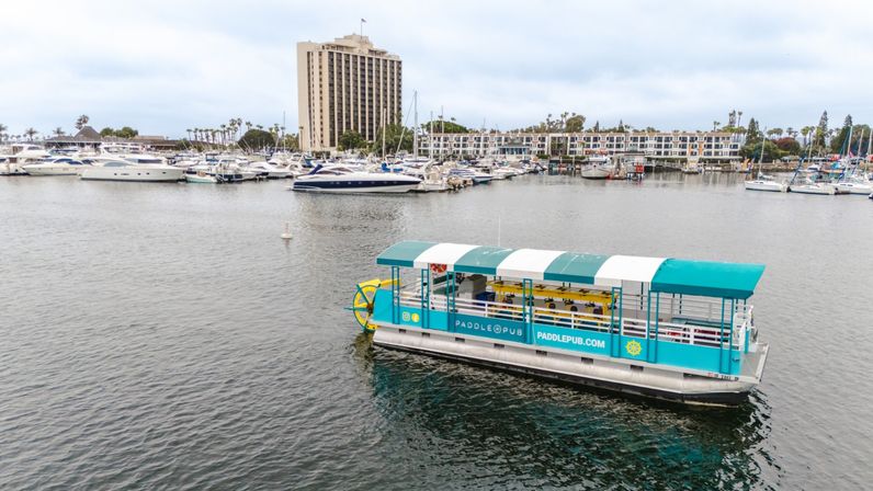 Turquoise-and-white paddle-wheel party boat cruising a coastal marina with moored yachts, waterfront condos, palm trees and a tall hotel under a cloudy sky
