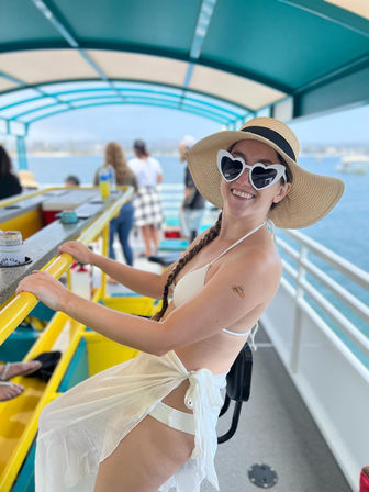 Smiling woman in a white bikini, sheer sarong, wide straw sun hat and heart-shaped sunglasses leaning on a yellow railing aboard a covered boat with turquoise canopy and ocean view on a sunny coastal cruise