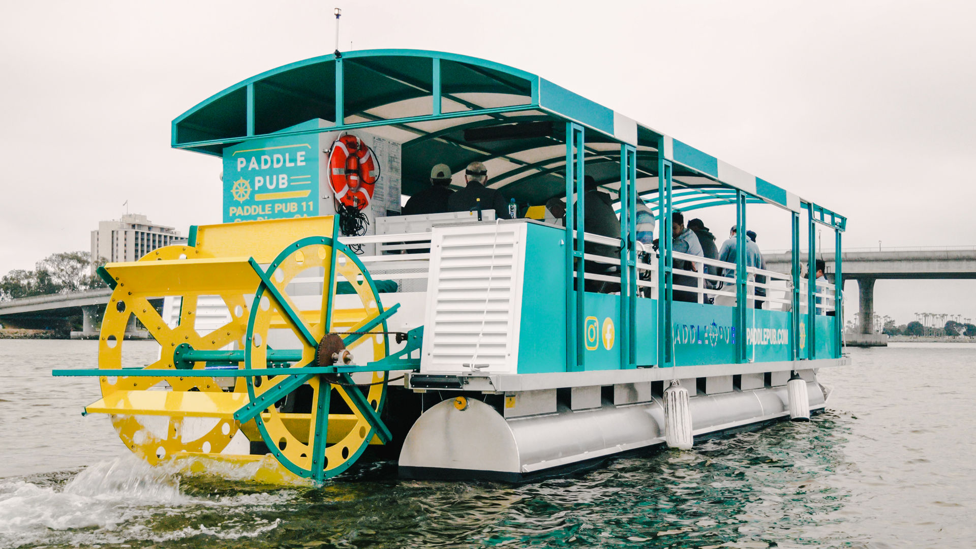 Teal and yellow paddlewheel party boat with passengers cruising on a calm river past a concrete bridge and waterfront buildings under an overcast sky