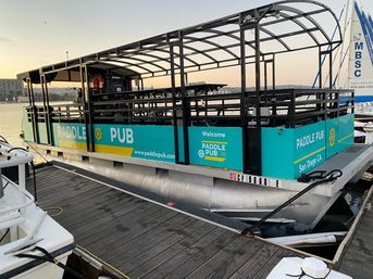 Turquoise party pontoon with covered open deck and bench seating docked at a San Diego marina at sunset, ready for a harbor cruise.
