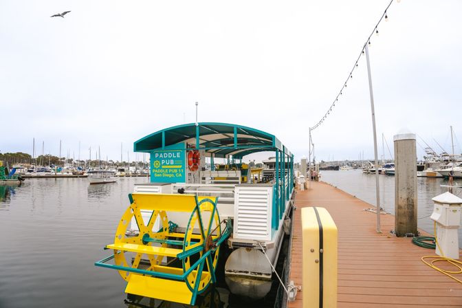 Bright yellow-and-teal paddlewheel boat docked at a San Diego marina beside a wooden pier with string lights and a seagull overhead.