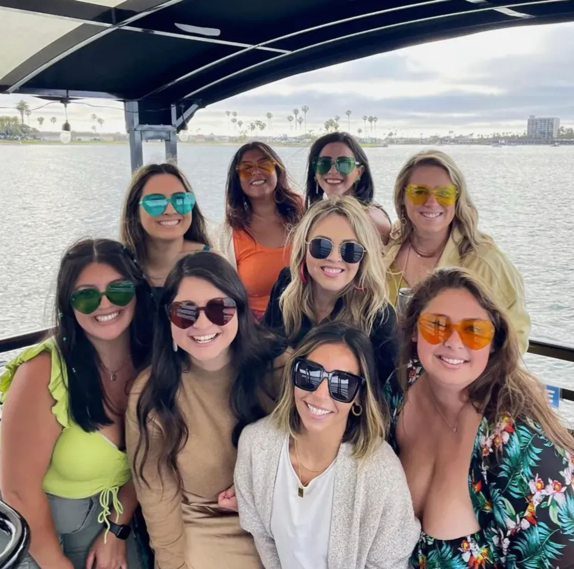 Nine friends in colorful sunglasses smiling together on a covered boat deck with a palm‑lined coastal bay and cloudy sky in the background.