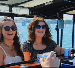 Two friends in sunglasses laughing on a sunny coastal boat bar, holding canned drinks with blue harbor water and a distant shoreline in the background.