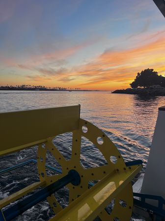 Yellow paddle wheel on a boat in the foreground with calm harbor waters reflecting a vibrant orange-pink sunset and a distant palm-lined shoreline.