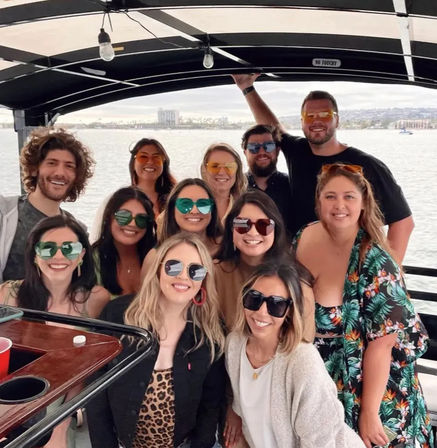 Smiling group of friends wearing colorful sunglasses on a covered party boat, posing by calm harbor waters with a coastal city skyline in the background.