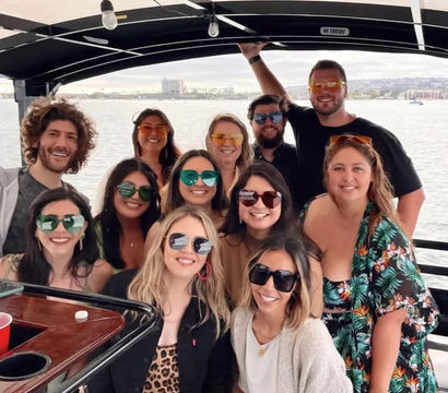 Smiling group of friends wearing colorful sunglasses on a covered party boat, posing by calm harbor waters with a coastal city skyline in the background.