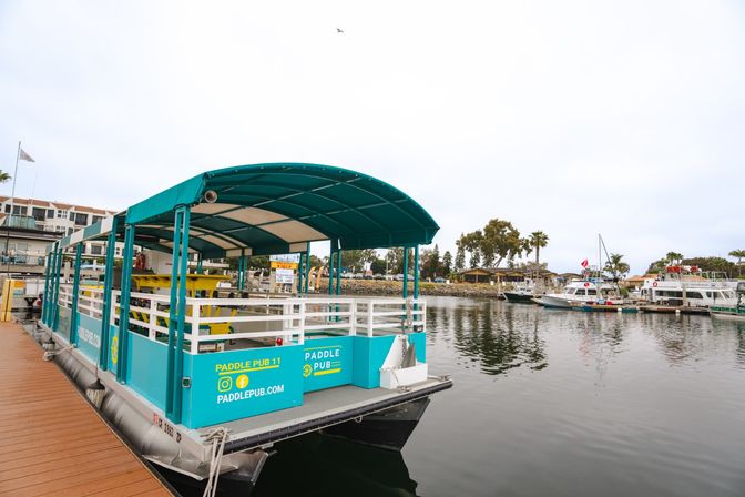 Turquoise covered party pontoon docked at a calm marina pier with other moored boats, palm trees along the shoreline and an overcast sky.