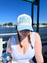Person on a sunny marina boat wearing a white and light-blue cap that reads 'MARRIAGE MATERIAL', white tank top and braids, holding a mirrored disco-ball tumbler with sailboats and blue sky in the background.