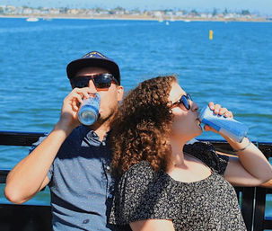 Two friends in sunglasses on a sunny boat ride in a coastal bay, leaning against the railing and sipping canned drinks with shoreline and palm trees in the background.