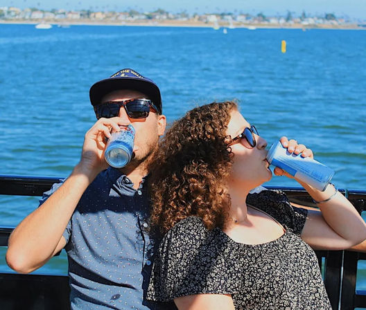 Two friends in sunglasses on a sunny boat ride in a coastal bay, leaning against the railing and sipping canned drinks with shoreline and palm trees in the background.