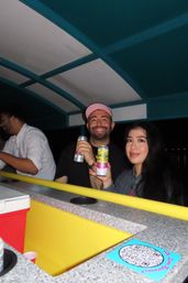 Two friends smiling on a nighttime boat bar, holding canned hard seltzers at a bright yellow counter under a teal canopy.