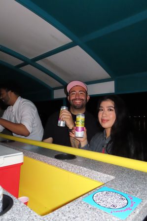 Two friends smiling on a nighttime boat bar, holding canned hard seltzers at a bright yellow counter under a teal canopy.
