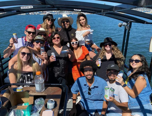 Smiling group of friends at a sunny boat party on a coastal marina, wearing sunglasses and summer hats while holding canned drinks.