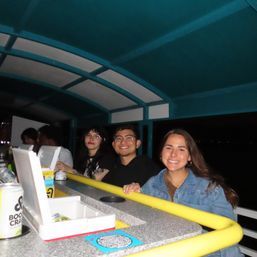 Three smiling friends leaning on a yellow railing at a covered boat bar at night, standing at a counter with canned drinks and takeout containers