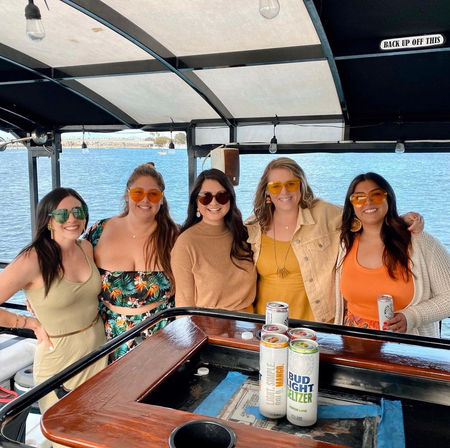 Five friends in colorful sunglasses smiling at a covered boat bar with canned seltzers, sunny blue harbor water and distant shoreline in the background