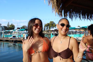 Two smiling friends in bikinis and sunglasses at a sunny marina dock with turquoise boat slips, string lights, a thatched canopy and calm water reflections
