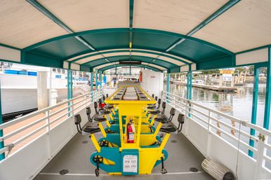 Covered pedal-powered party boat with bright yellow central pedal station and black seats docked at a marina harbor with calm water and nearby boats