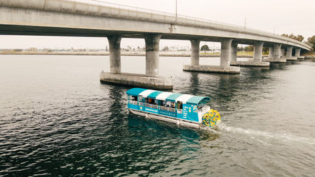 Blue-and-white covered paddleboat with a bright yellow rear paddle wheel cruising under a concrete bridge across a calm coastal bay