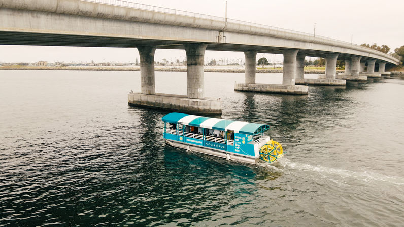 Blue-and-white covered paddleboat with a bright yellow rear paddle wheel cruising under a concrete bridge across a calm coastal bay