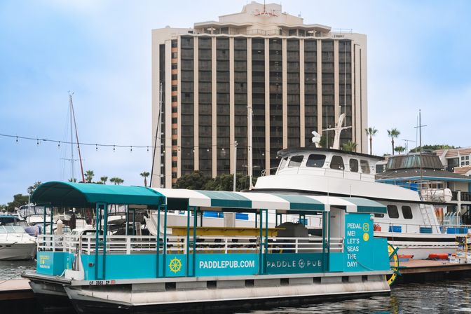 Teal pedal-powered party boat docked at a coastal marina with a white yacht, palm trees and a tall waterfront hotel in the background.