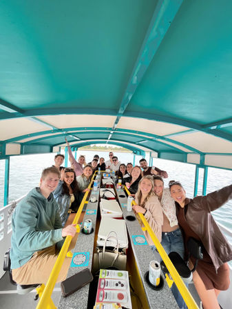 Smiling group of people on a covered pontoon boat lake cruise, gathered around a central bar under a teal canopy with drinks and coolers visible.