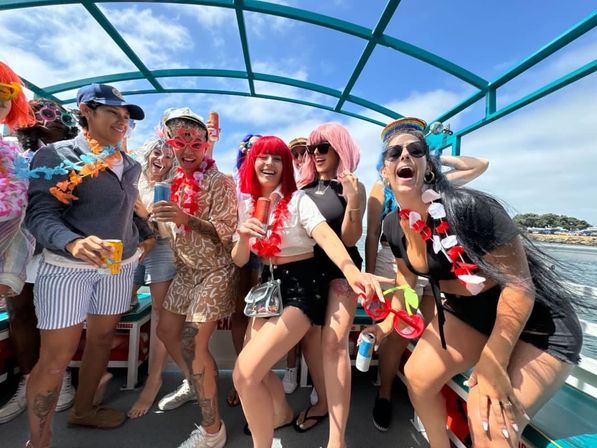 Group of friends at a sunny coastal boat party, wearing colorful wigs, leis and sunglasses, laughing and holding canned drinks under a turquoise canopy with ocean and rocky shoreline in the background.