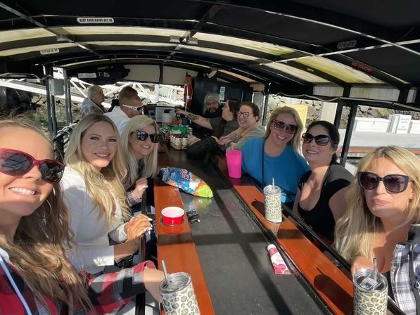 Group of women smiling around a wooden bar inside a covered party boat at a sunny marina with drinks, sunglasses and snacks