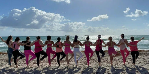 Group of people in pink activewear linked arm‑in‑arm doing tree pose on a sunny sandy beach, facing the sparkling ocean and fluffy clouds on the horizon