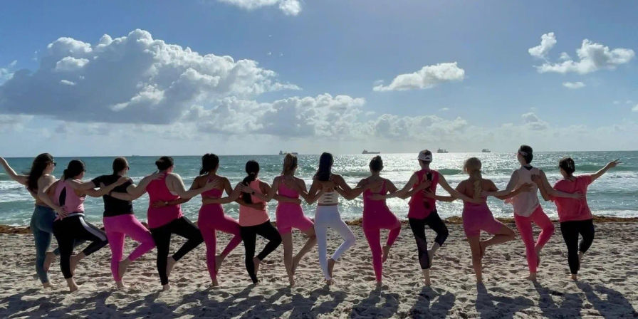 Group of people in pink activewear linked arm‑in‑arm doing tree pose on a sunny sandy beach, facing the sparkling ocean and fluffy clouds on the horizon