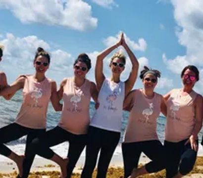 Seven friends in matching pink and white tank tops and black leggings strike tree-pose yoga on a sunny sandy oceanfront beach under a blue sky with fluffy clouds.