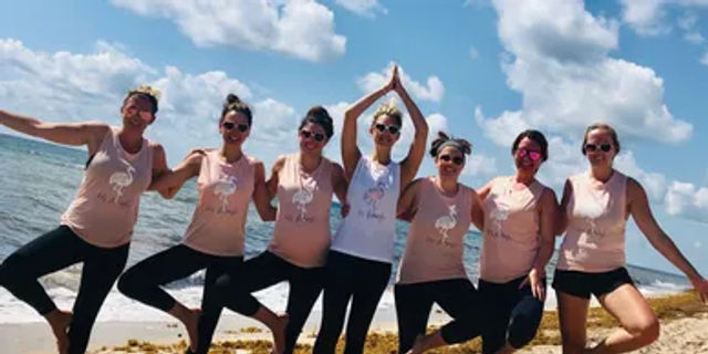 Seven friends in matching pink and white tank tops and black leggings strike tree-pose yoga on a sunny sandy oceanfront beach under a blue sky with fluffy clouds.