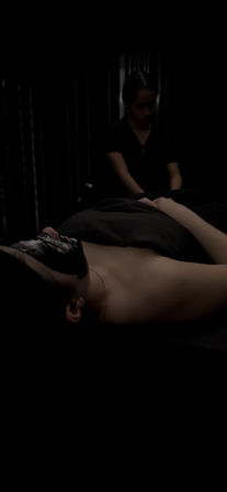 Relaxing spa scene: reclining client with a black facial mask and head wrap, covered by a dark sheet while an esthetician works in a dimly lit treatment room.