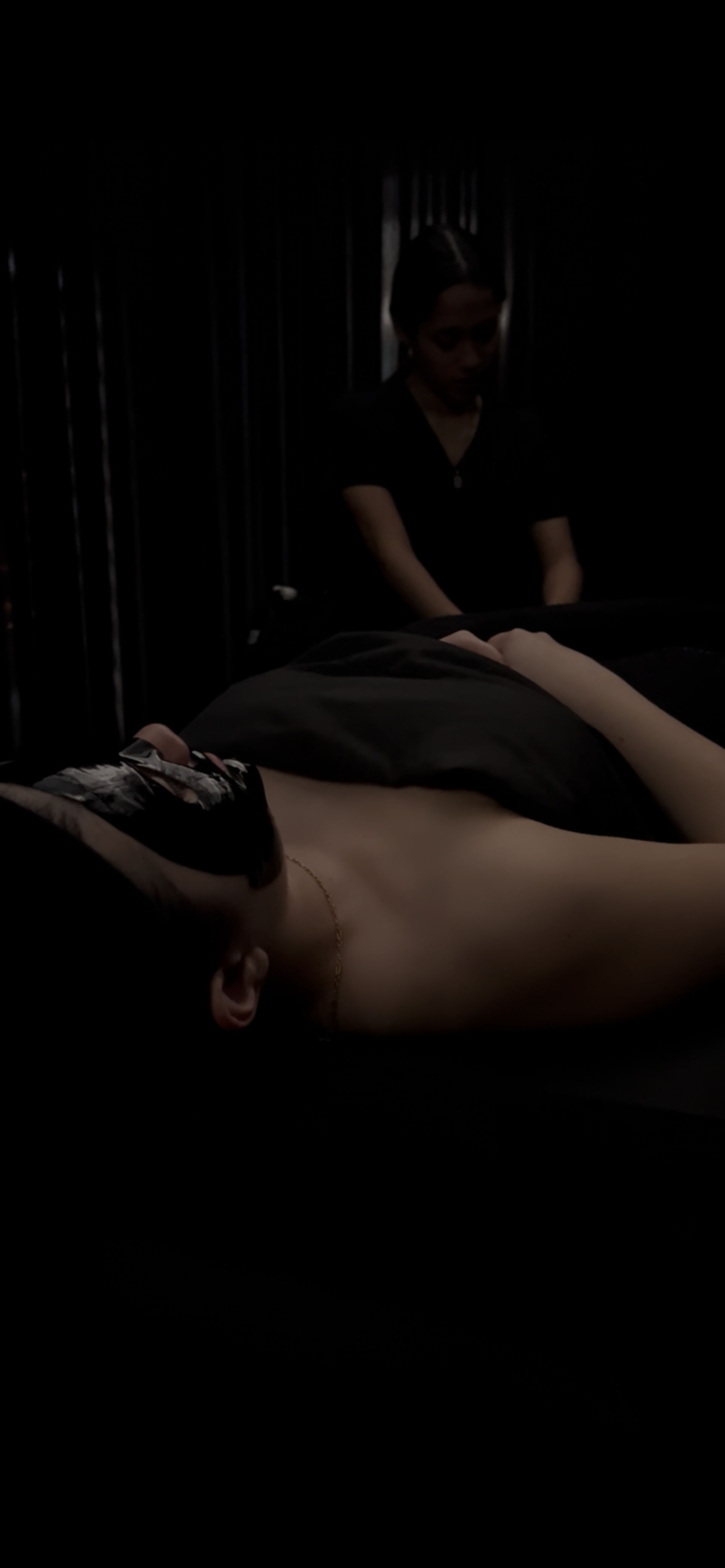Relaxing spa scene: reclining client with a black facial mask and head wrap, covered by a dark sheet while an esthetician works in a dimly lit treatment room.