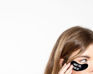 Close-up of a person applying a black under-eye gel patch printed "WAKE THE F*** UP" on a white background — playful skincare/beauty morning routine.