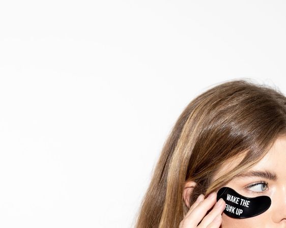 Close-up of a person applying a black under-eye gel patch printed "WAKE THE F*** UP" on a white background — playful skincare/beauty morning routine.