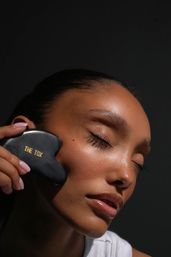 Close-up of a woman with dewy skin and closed eyes massaging her cheek with a black heart-shaped gua sha tool stamped "THE TOX" against a dark background