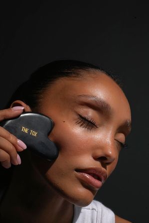 Close-up of a woman with dewy skin and closed eyes massaging her cheek with a black heart-shaped gua sha tool stamped "THE TOX" against a dark background
