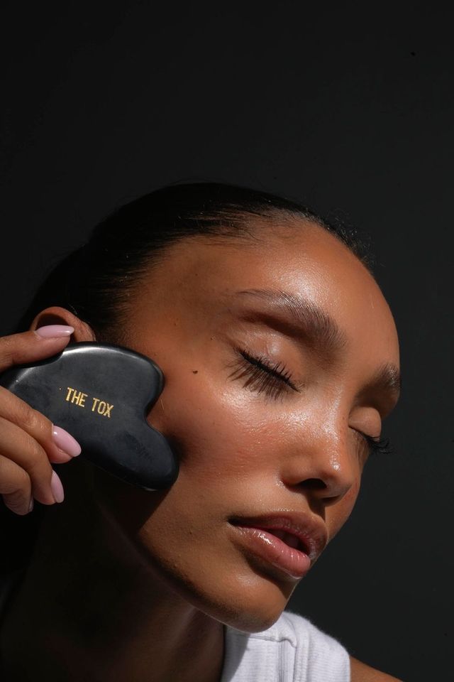 Close-up of a woman with dewy skin and closed eyes massaging her cheek with a black heart-shaped gua sha tool stamped "THE TOX" against a dark background